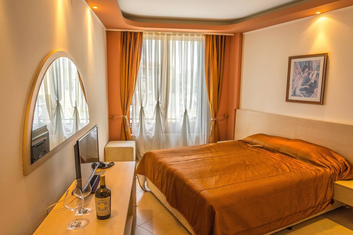 Une chambre d’hôtel avec un lit, un bureau sur lequel se trouve une bouteille de vin et des verres, un miroir et des rideaux.
