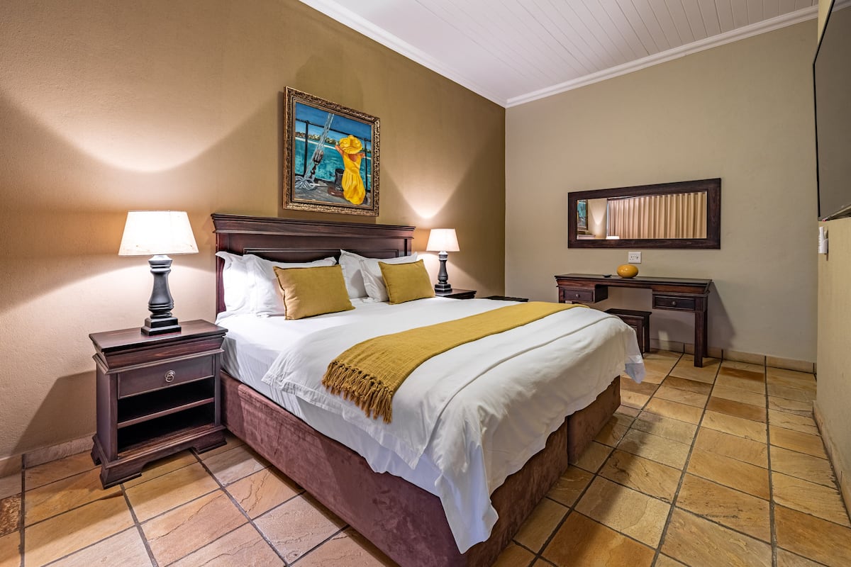 Een net opgemaakt bed met een deken in witte en mosterdtinten, twee nachtlampen, een nachtkastje, een houten hoofdbord, een schilderij boven het bed, een klein bureau met een spiegel en een betegelde vloer.