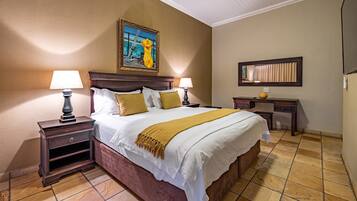 A neatly made bed with a white and mustard blanket, two bedside lamps, a nightstand, a wooden headboard, a painting above the bed, a small desk with a mirror, and a tiled floor.