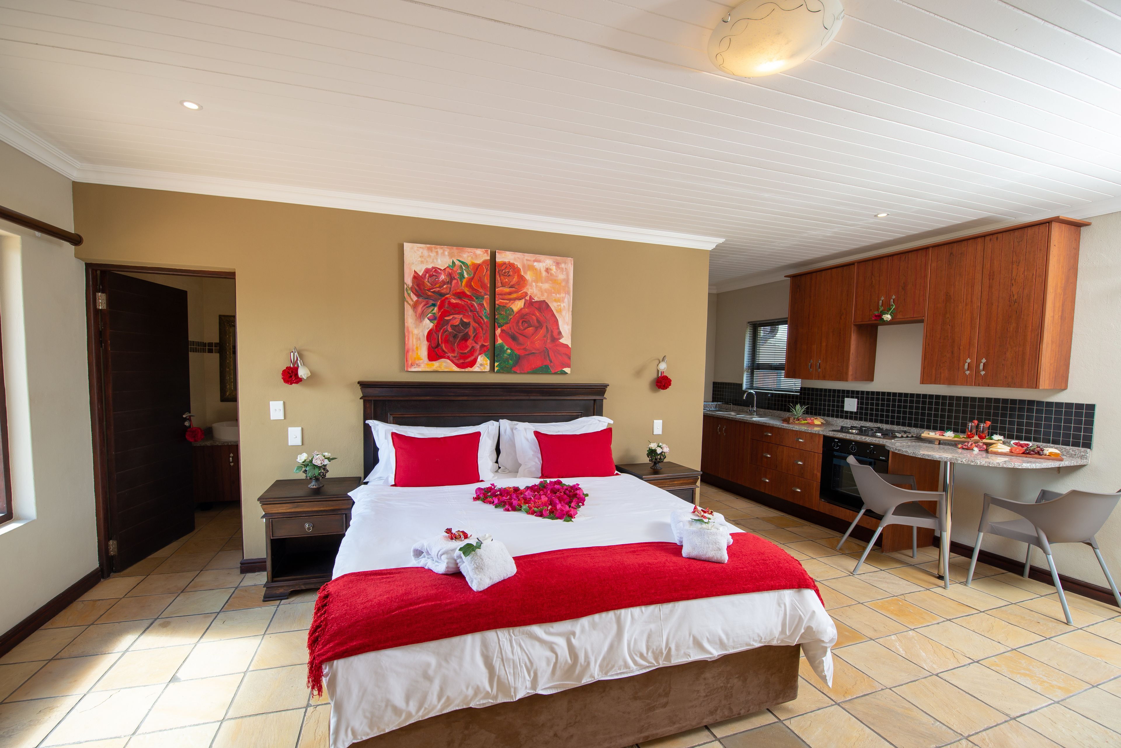 A hotel room with a large bed, a painting of red roses, a small table with a vase, and a kitchenette area.