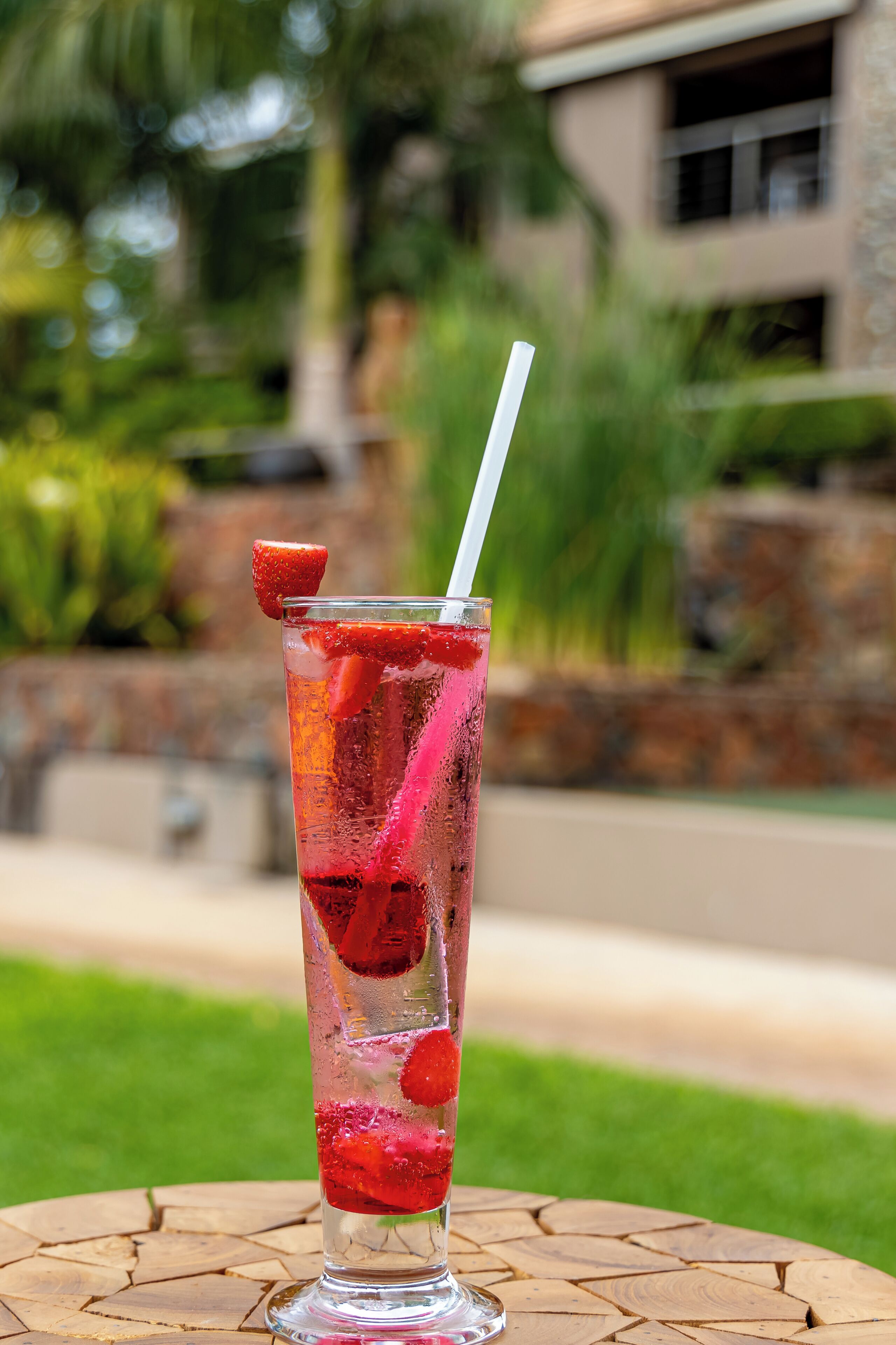 Un vaso alto con pajita, lleno de una bebida roja y adornado con fruta, sobre una mesa de madera redonda al aire libre.