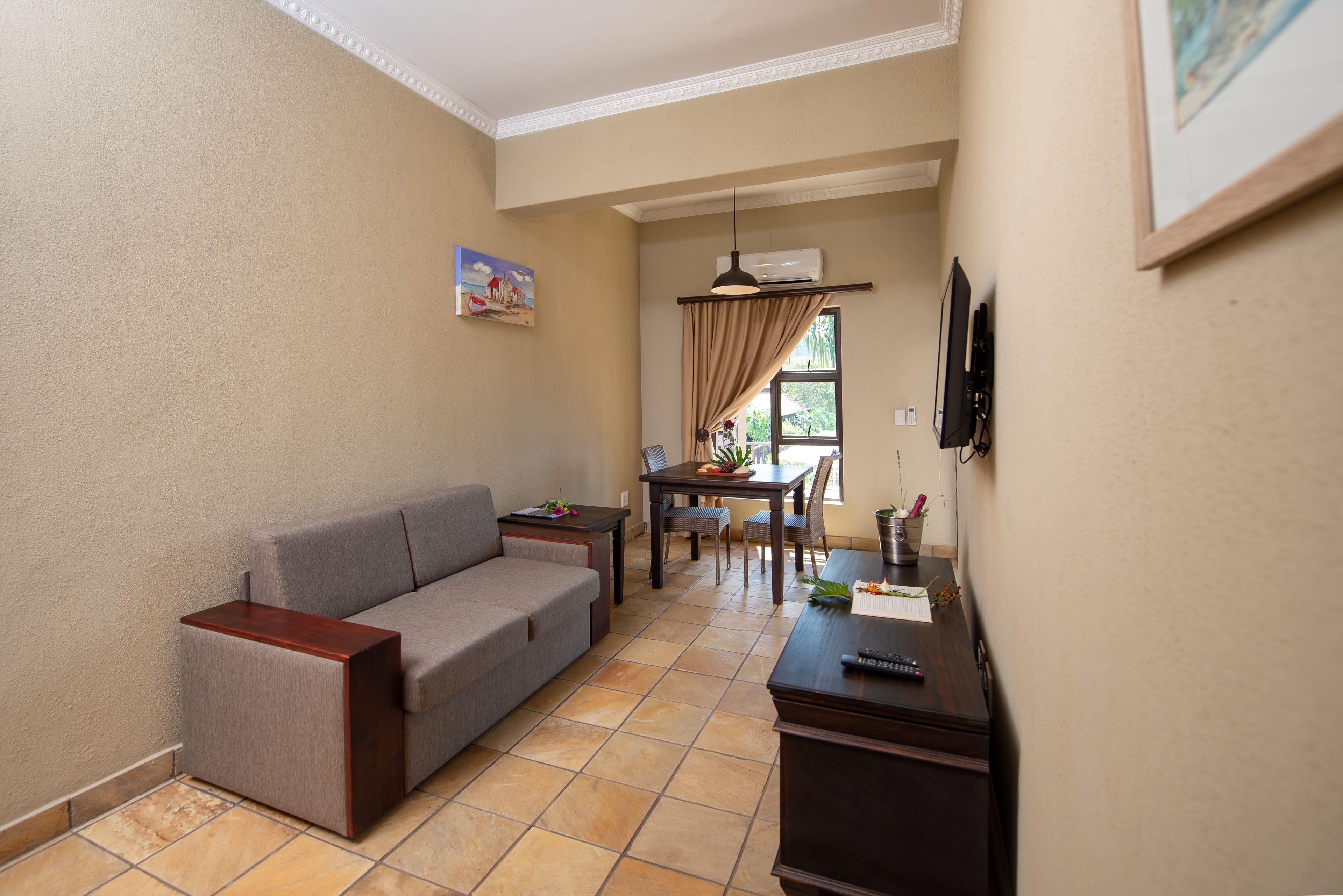 A living room with a grey sofa, a wooden side table, a dining table with chairs, and a TV mounted on the wall.