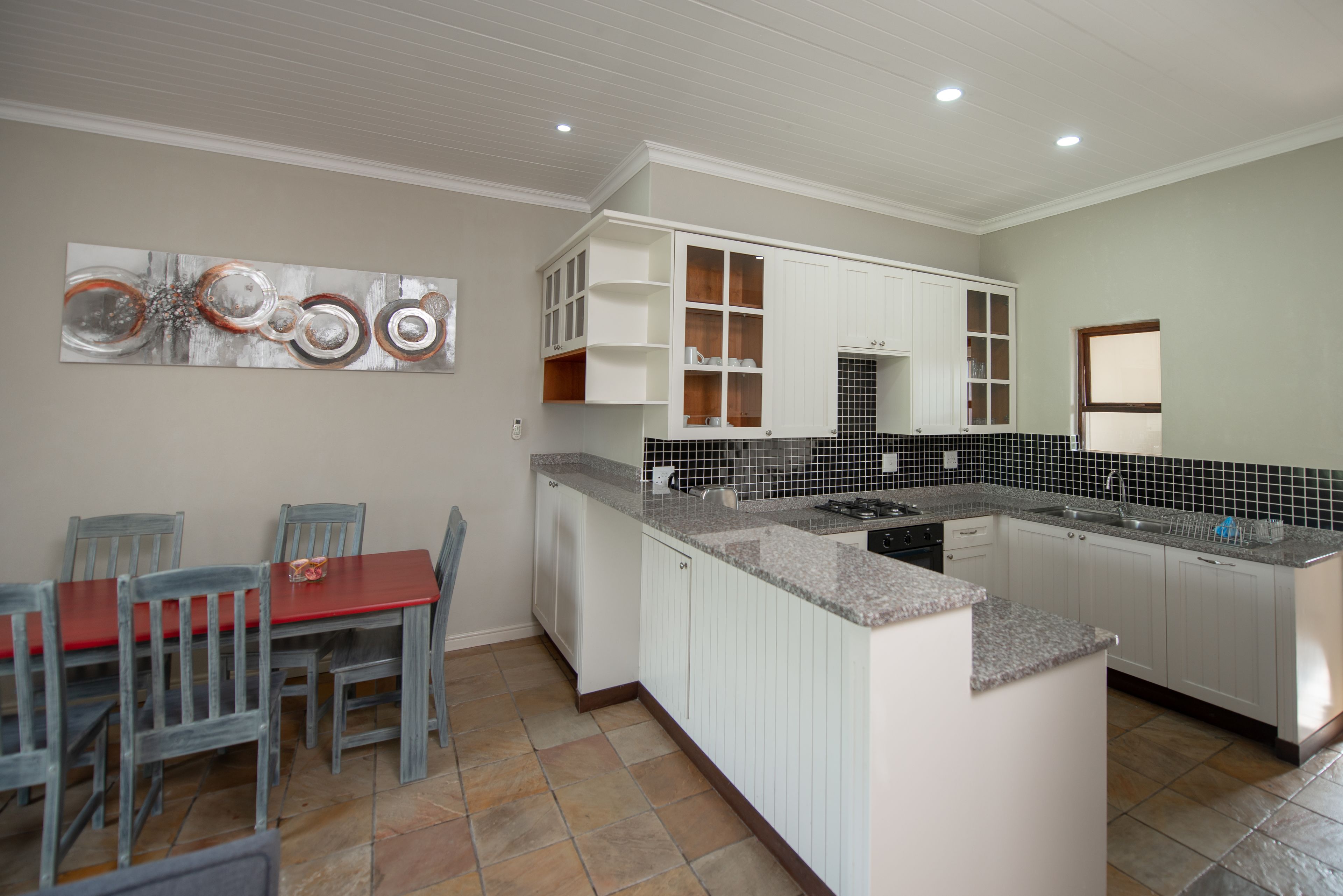 Una cocina con armarios blancos, encimera de granito y comedor con mesa roja y sillas grises.