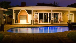 A covered outdoor pool area with a brick patio, a covered seating area with chairs, and a building with a brick fireplace. - McAllisters on 8th (Durban)