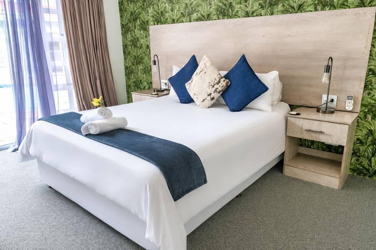 A hotel room with a bed, a nightstand, a lamp, and a view of greenery outside.
