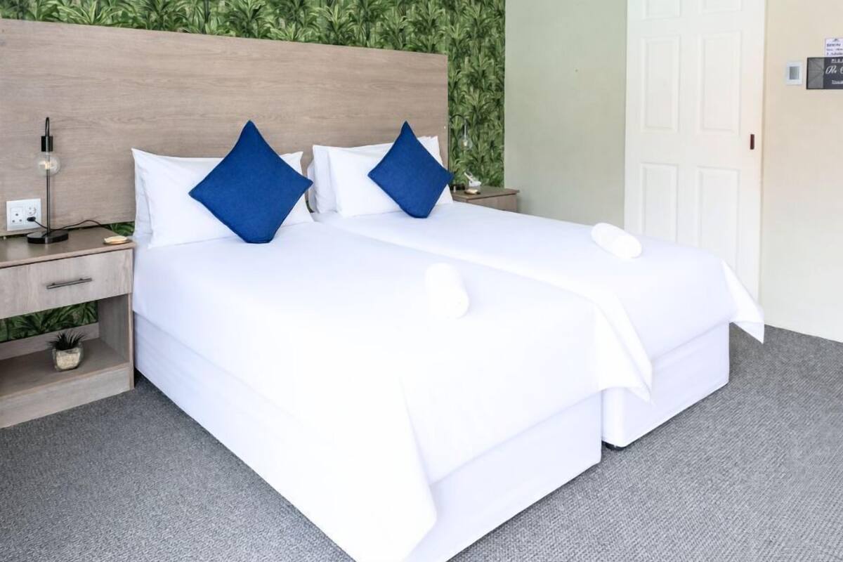 A double bed with white bedding and two blue pillows, a bedside table with a lamp, a small plant, and a door with a sign.