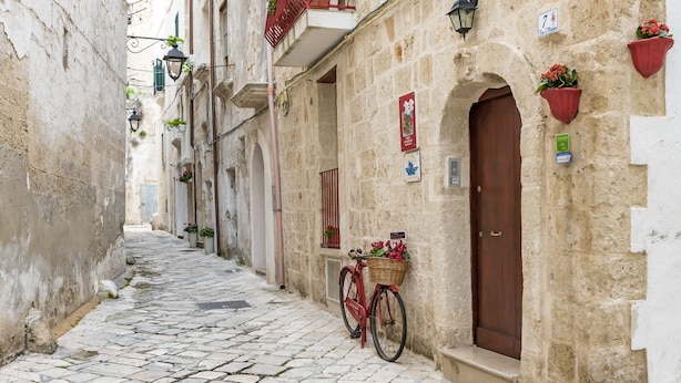 Una estrecha calle empedrada con una bicicleta roja estacionada junto a un edificio con una puerta marrón y macetas con flores.
