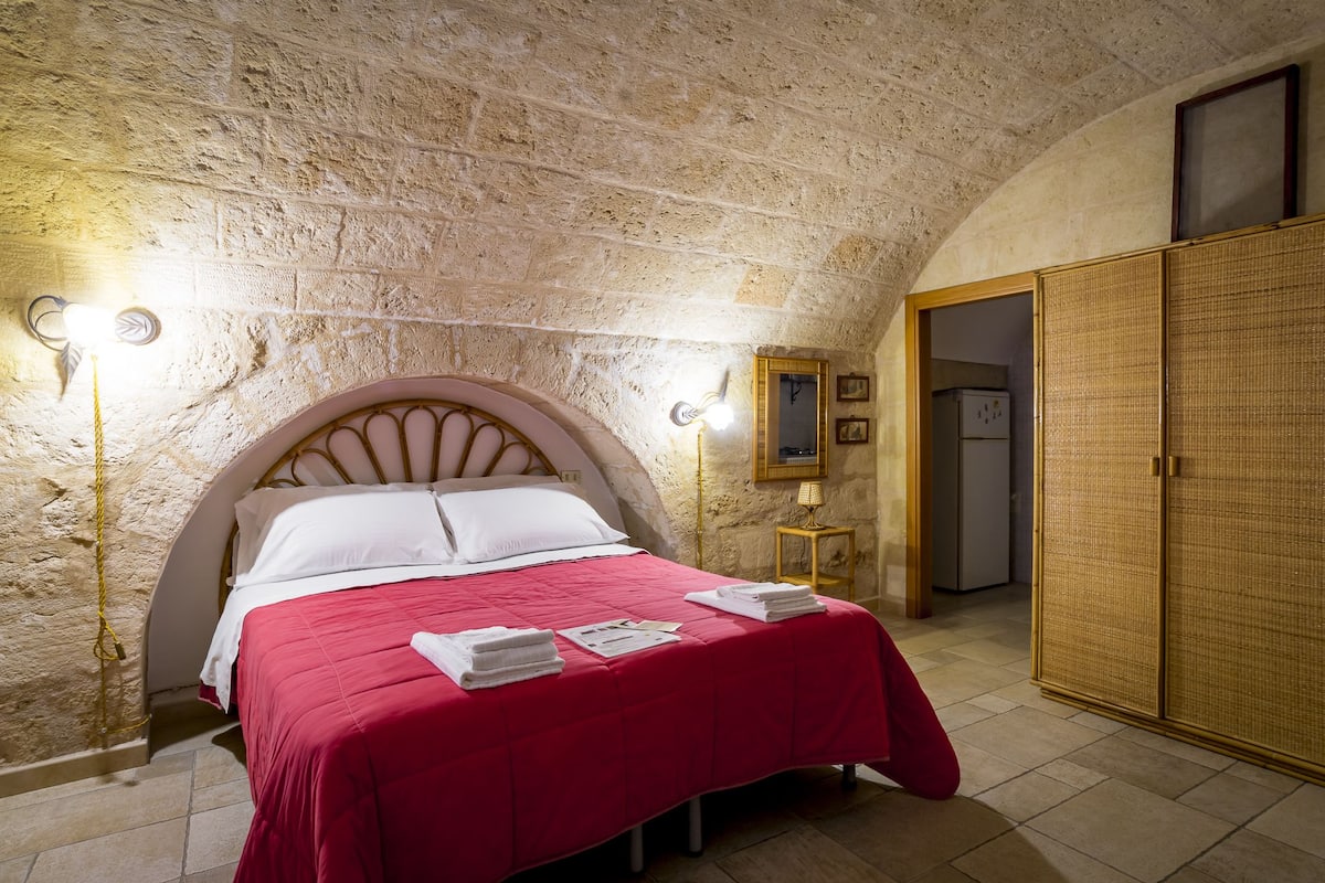 Una habitación con pared de piedra, cama con colcha roja, dos lámparas de noche, armario de madera y una mesita con lámpara.