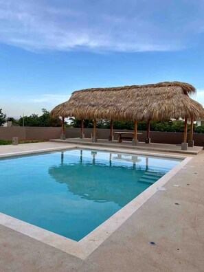 A swimming pool with a thatched roof structure, surrounded by a concrete deck.