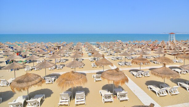 A beach with rows of white lounge chairs and thatched umbrellas, a lifeguard tower, and a clear blue sky.