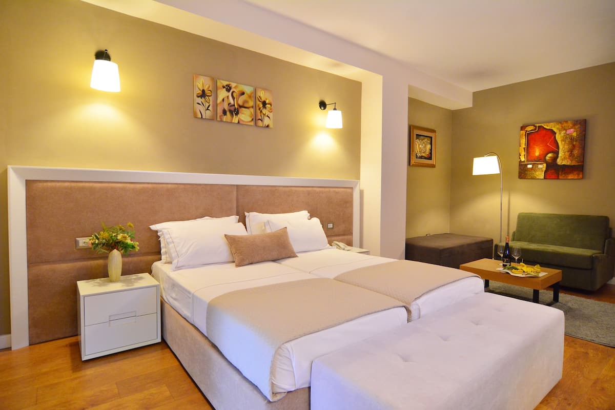 A hotel room with a large bed, bedside tables, a nightstand with a vase, a green sofa, and artwork on the walls.