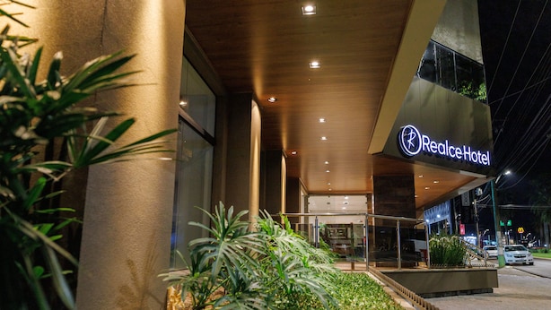 Exterior de un hotel con diseño moderno, destacando el letrero RealceHotel, una entrada bien iluminada y una zona ajardinada con plantas.