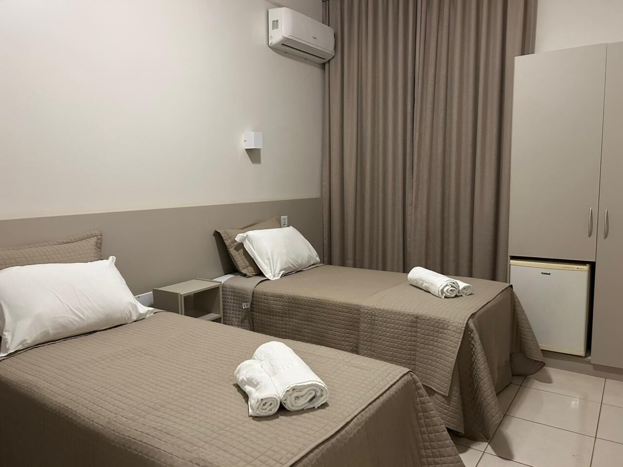 A hotel room with two beds, a small table, a wardrobe, and a mini-fridge.