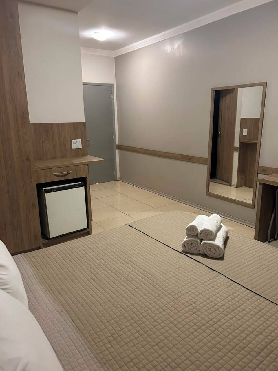 A hotel room with a bed, a small refrigerator, a mirror, and a pair of rolled towels on the floor.