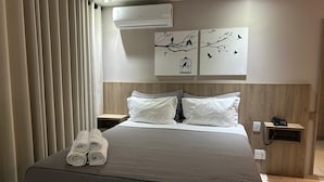 Una cama bien tendida con una colcha gris, almohadas blancas y toallas enrolladas al lado. Hay dos obras de arte colgadas en la pared que representan pájaros en las ramas.
