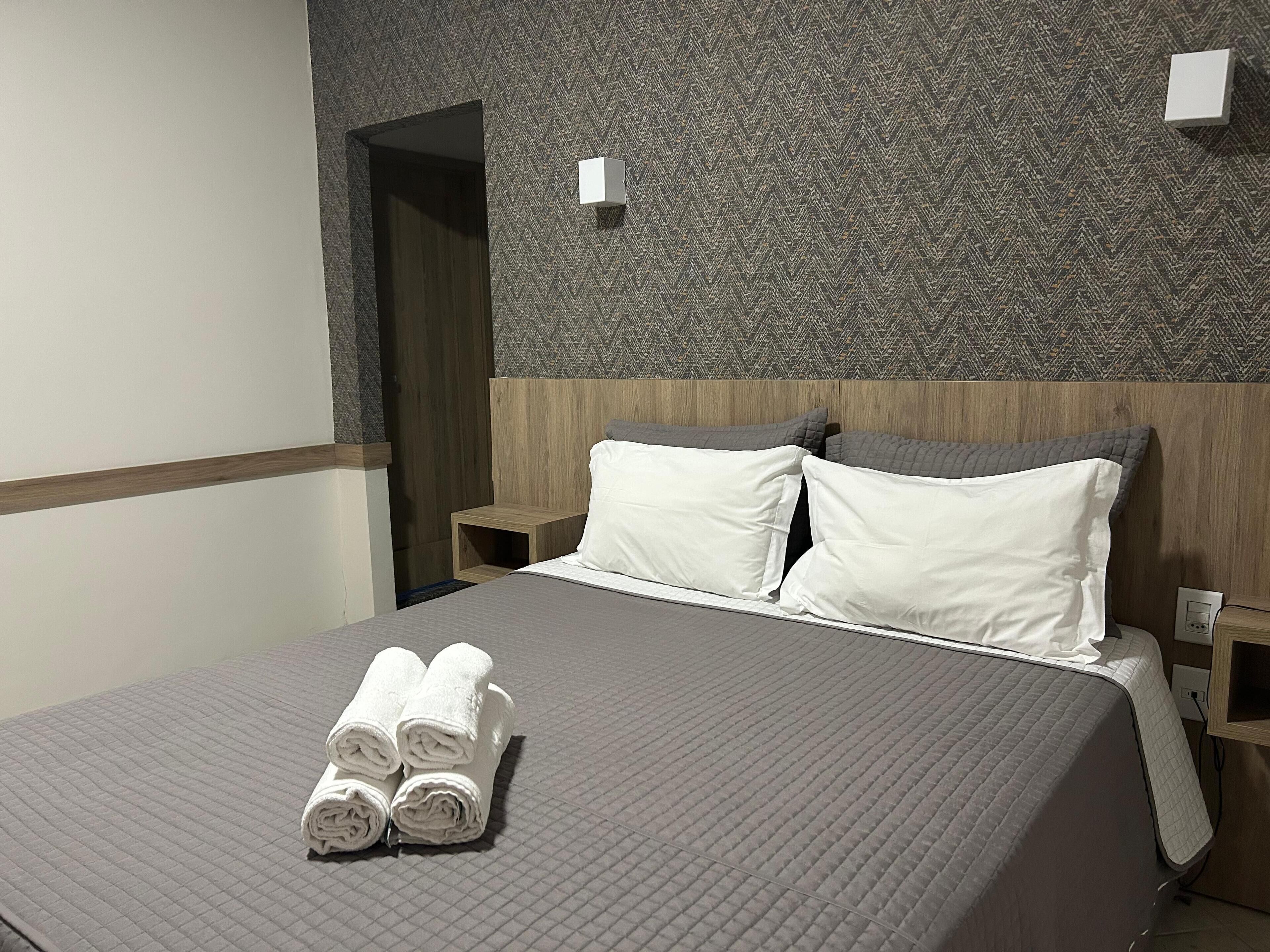 Una cama bien hecha con almohadas blancas y toallas enrolladas, un cabecero de madera y un patrón texturizado en la pared.