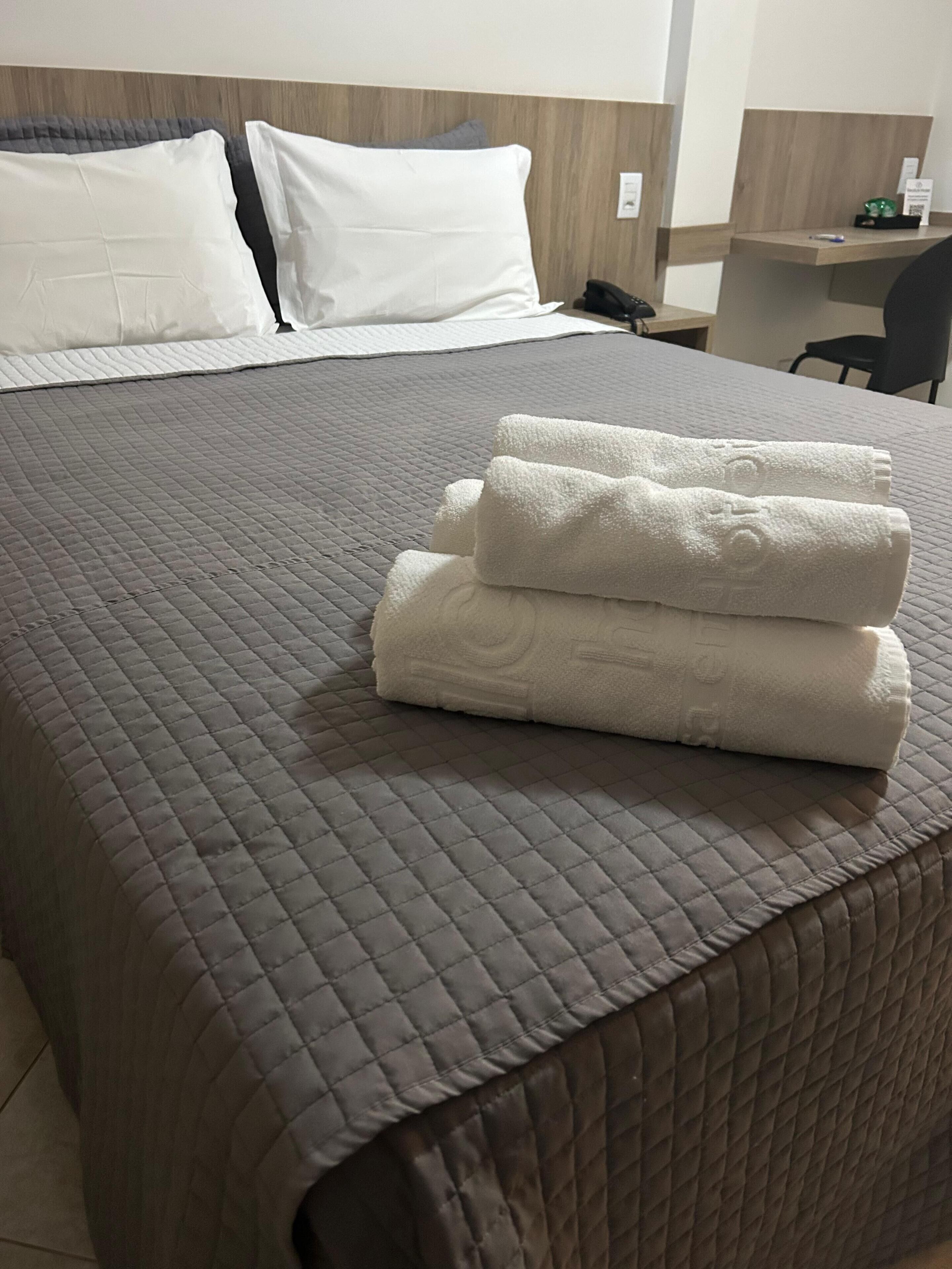 Una cama bien hecha con una colcha gris acolchada, dos almohadas blancas y una toalla blanca doblada sobre la cama.