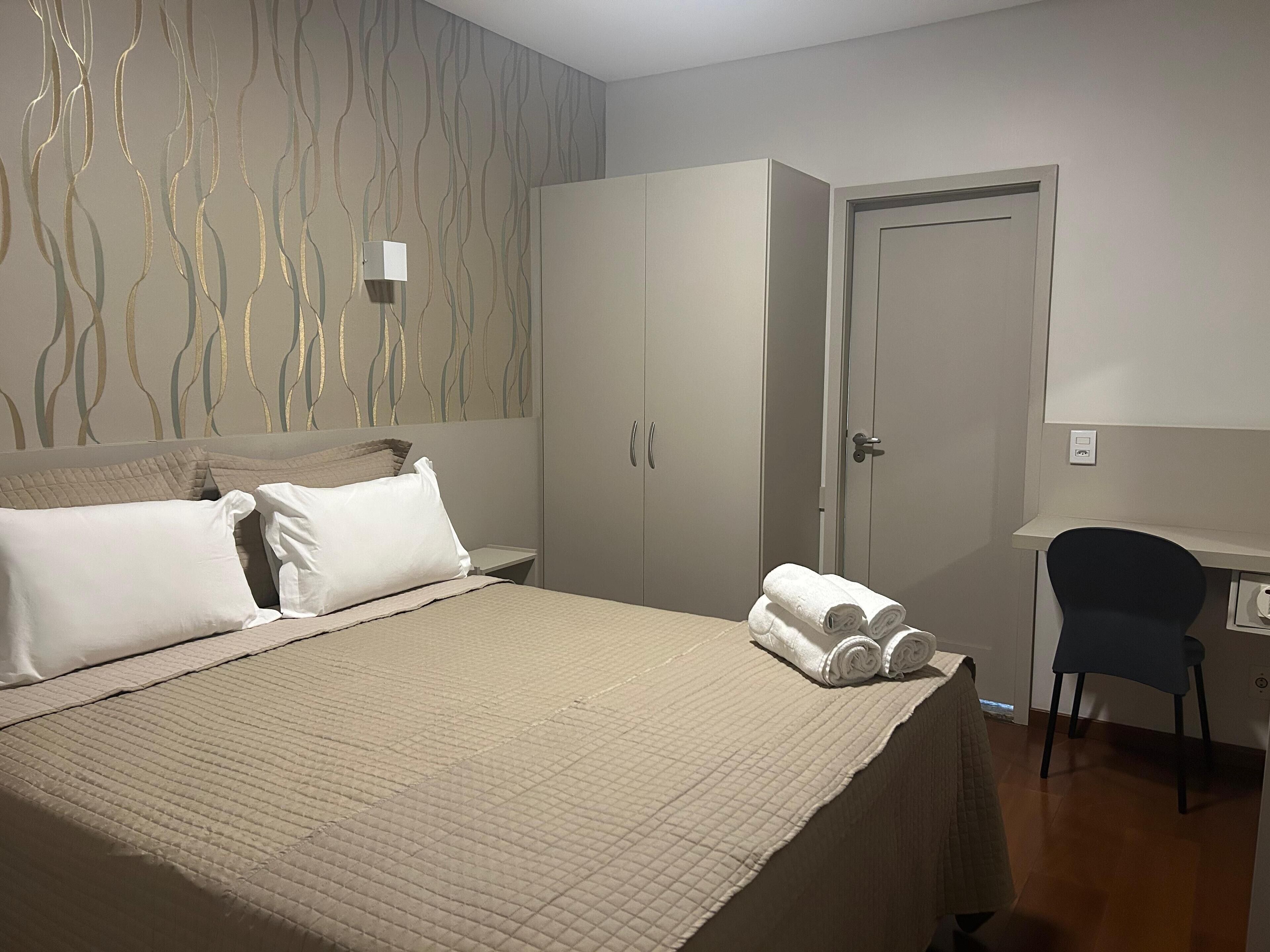A neatly made bed with a quilted cover, a desk with a chair, and a wardrobe with two doors.