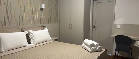 A neatly made bed with a quilted cover, a desk with a chair, and a wardrobe with two doors.