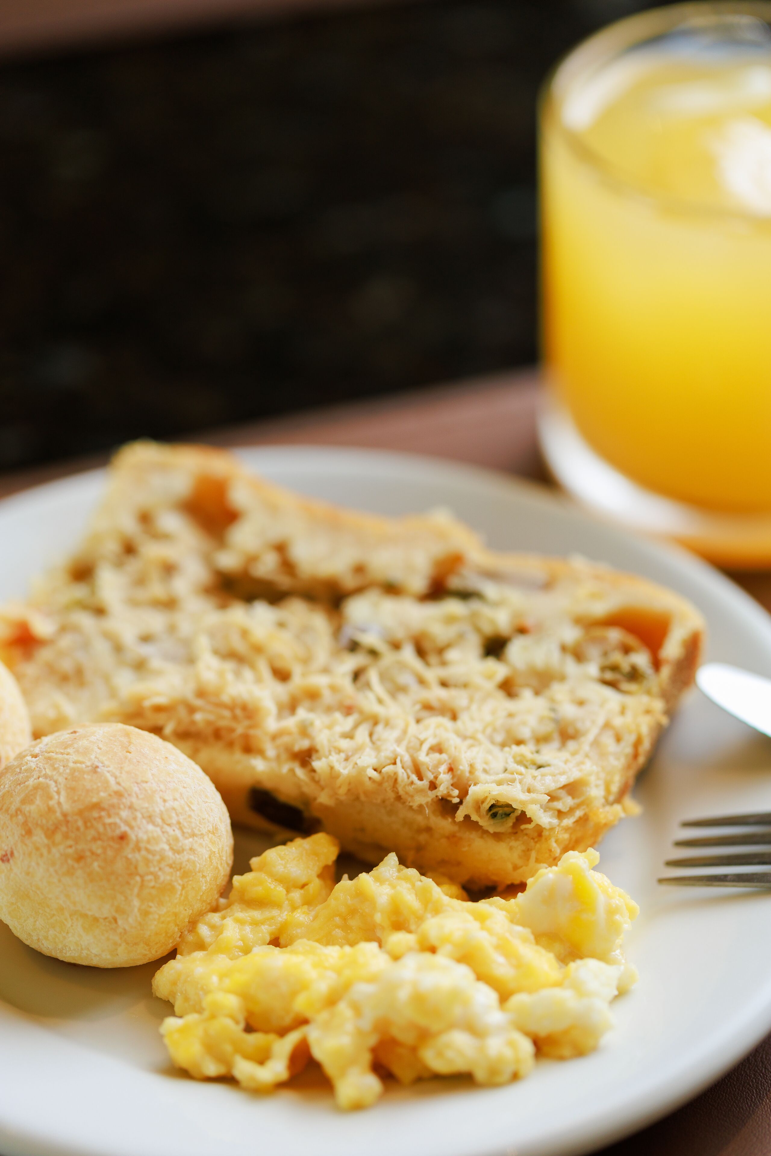 A plate with scrambled eggs, a slice of quiche, and a small round bread.