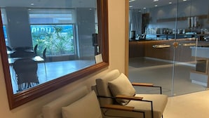 Un área de espera moderna con un ventanal grande, una puerta de vidrio que da a una cocina y una disposición de sofás y sillones.