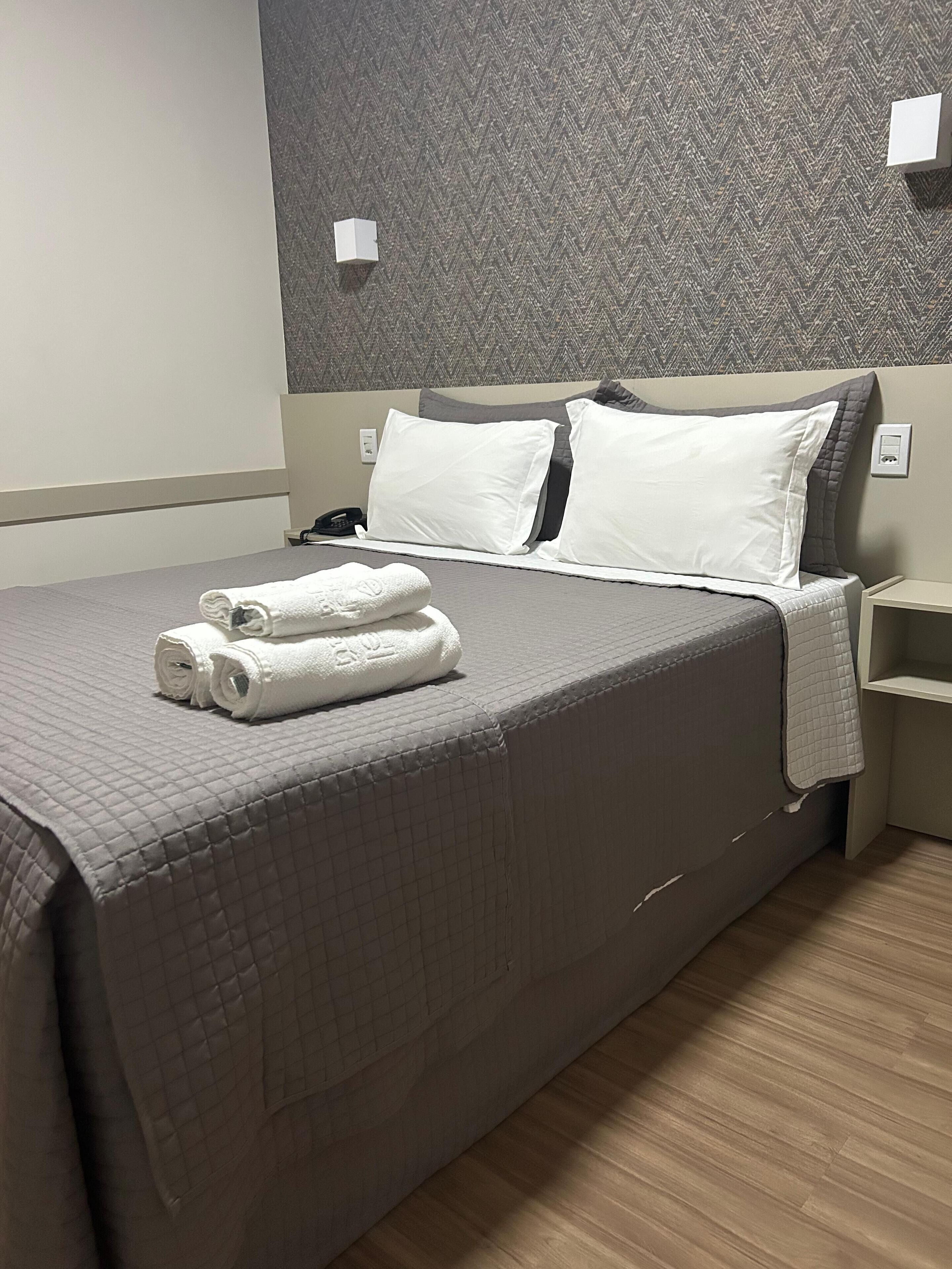 Una cama bien hecha con una colcha gris acolchada, dos almohadas blancas y dos toallas enrolladas en blanco.