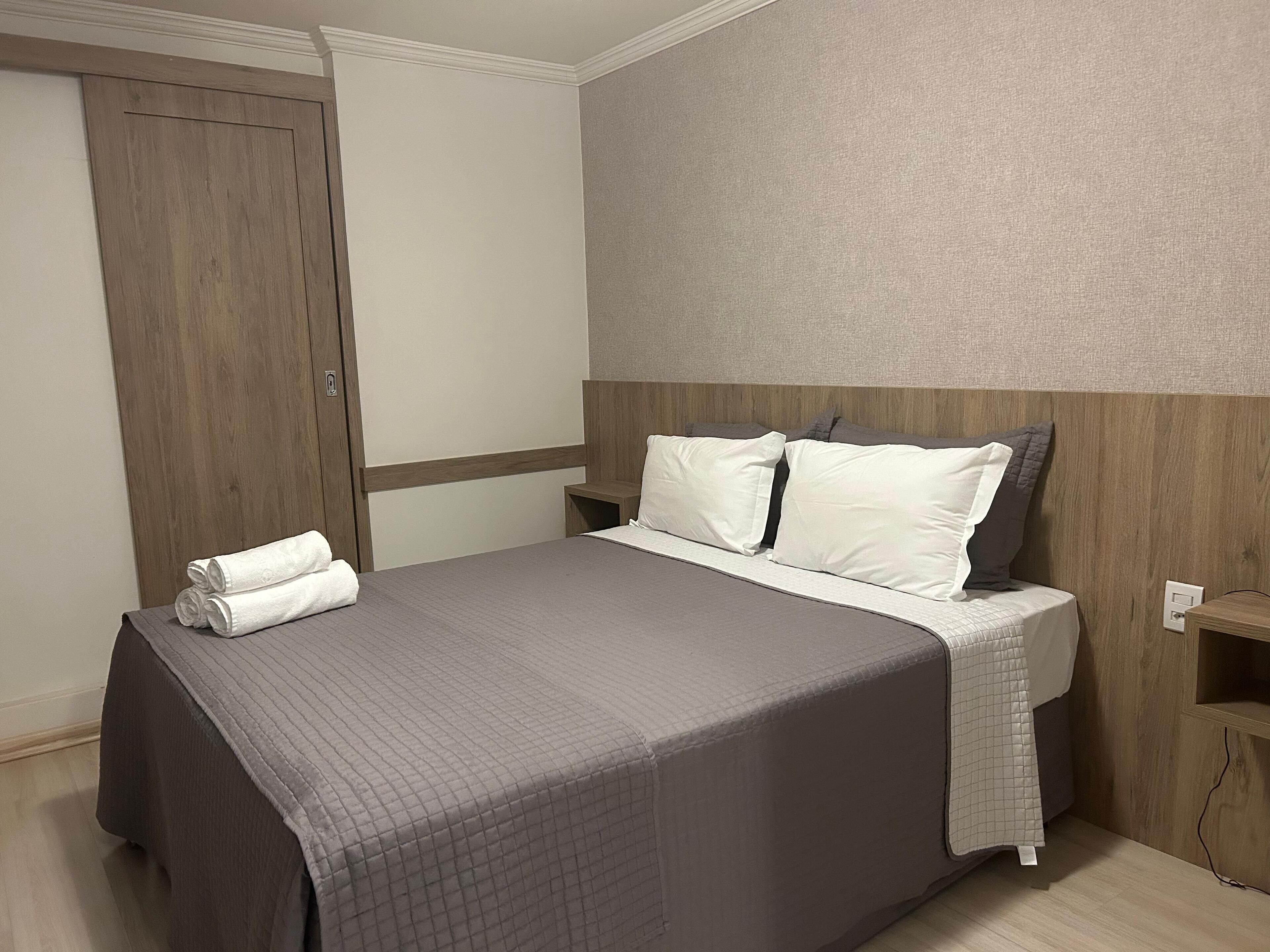 A neatly made bed with a gray quilt and white pillows, a wooden headboard, a wooden wardrobe, and a small wooden nightstand with a lamp.