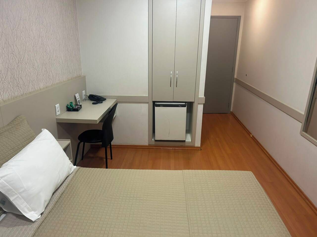 A compact hotel room with a bed, a desk with a chair, a small refrigerator, and a wardrobe.