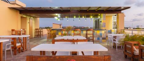 An outdoor patio with wooden tables and benches, a covered seating area, and a view of the cityscape.