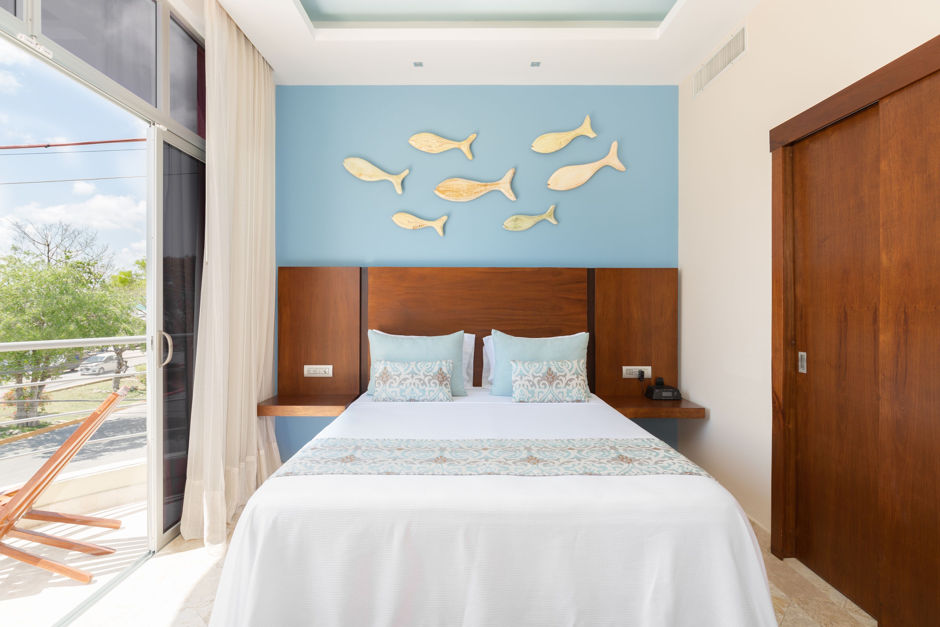 A hotel room with a bed, a wooden headboard, a wall with fish-shaped decorations, and a balcony with a view of the outside.