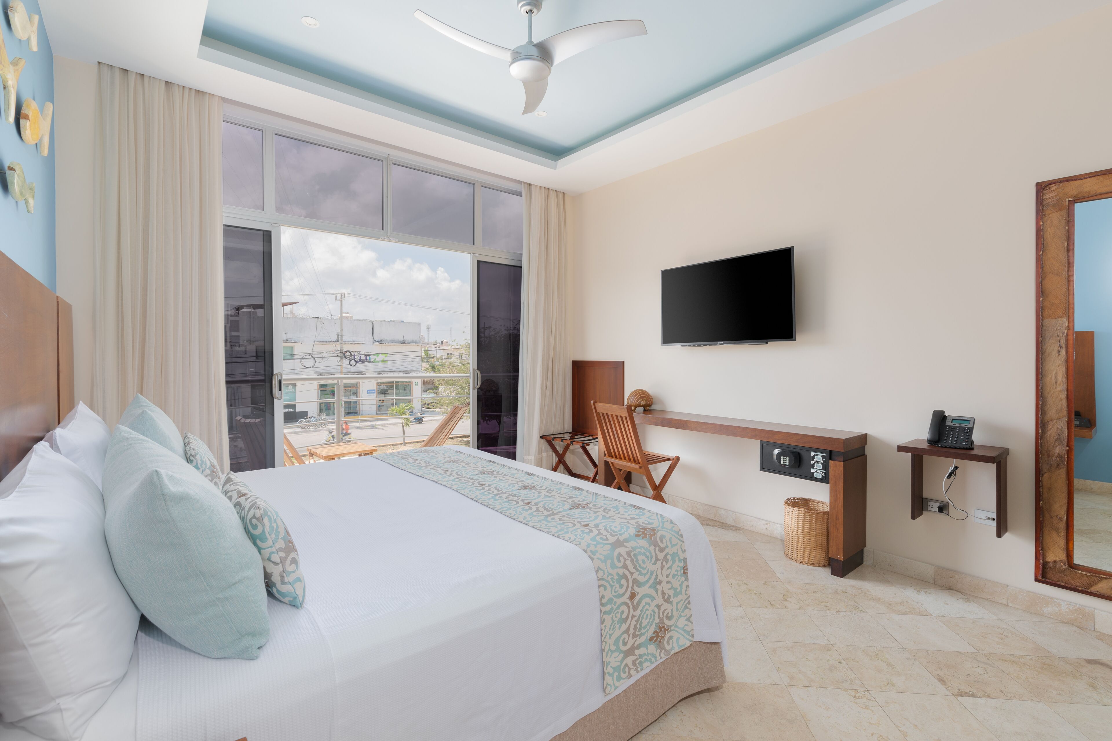 A hotel room with a large bed, a flat-screen TV mounted on the wall, a wooden chair, a small desk with a phone, and a view of the city through large windows.