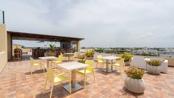 Una terraza en la azotea con sillas y mesas amarillas, asientos al aire libre y plantas en macetas.