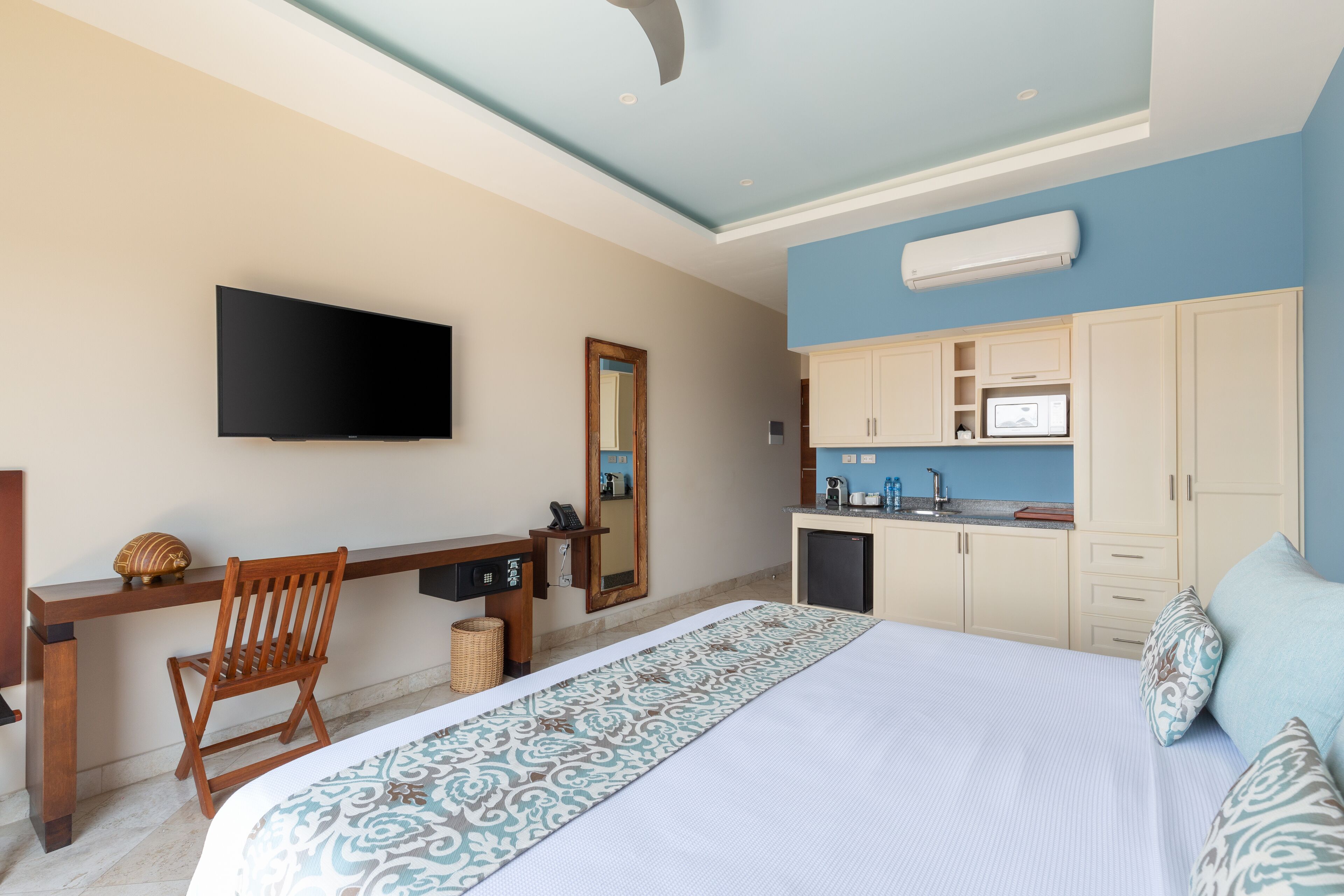 Een hotelkamer met een bed, een bureau, een televisie, een spiegel, een kitchenette en een plafondventilator.