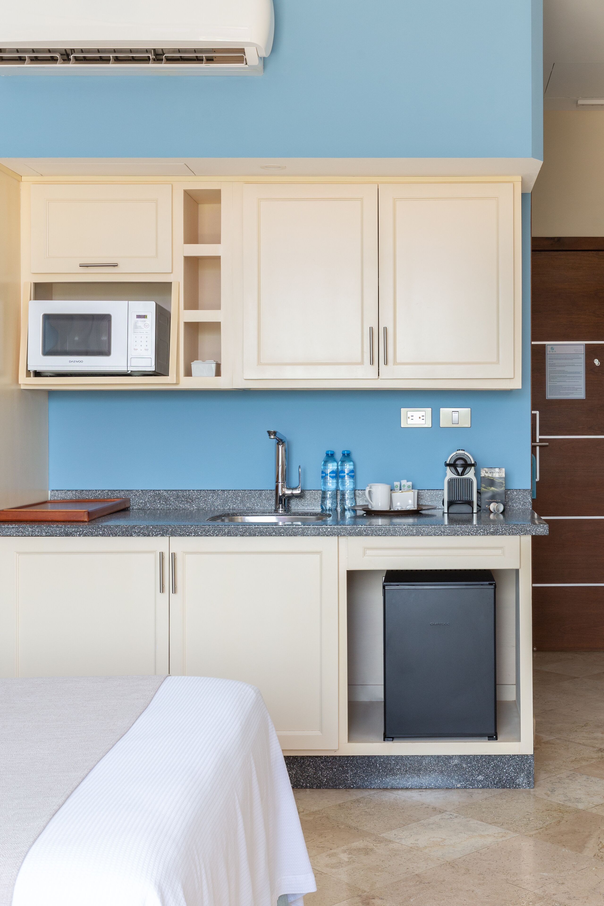 A compact hotel room with a kitchenette, a bed with a white bedspread, and a small refrigerator.