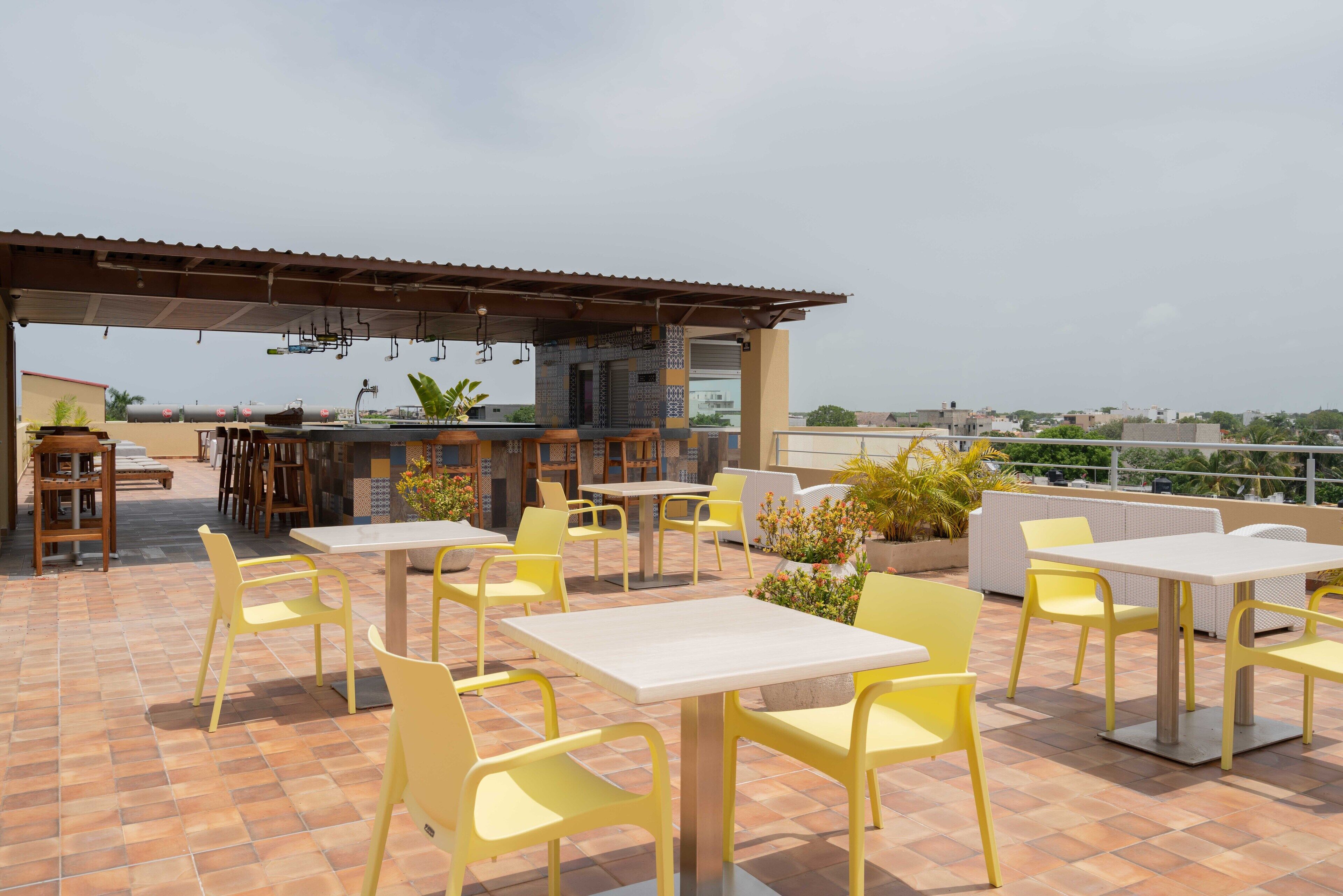 Un patio al aire libre con sillas y mesas amarillas, una zona de bar y vistas a la ciudad.