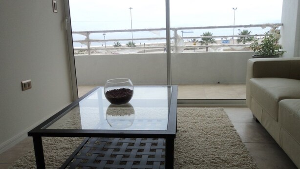 A living room with a glass coffee table, beige sofa, and a view of a beach and bridge. - Depto Claudia Peñuelas (Coquimbo)