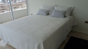 A bedroom with a large bed, two pillows, and a view of a construction site through a glass door. - Depto Claudia Peñuelas (Coquimbo)