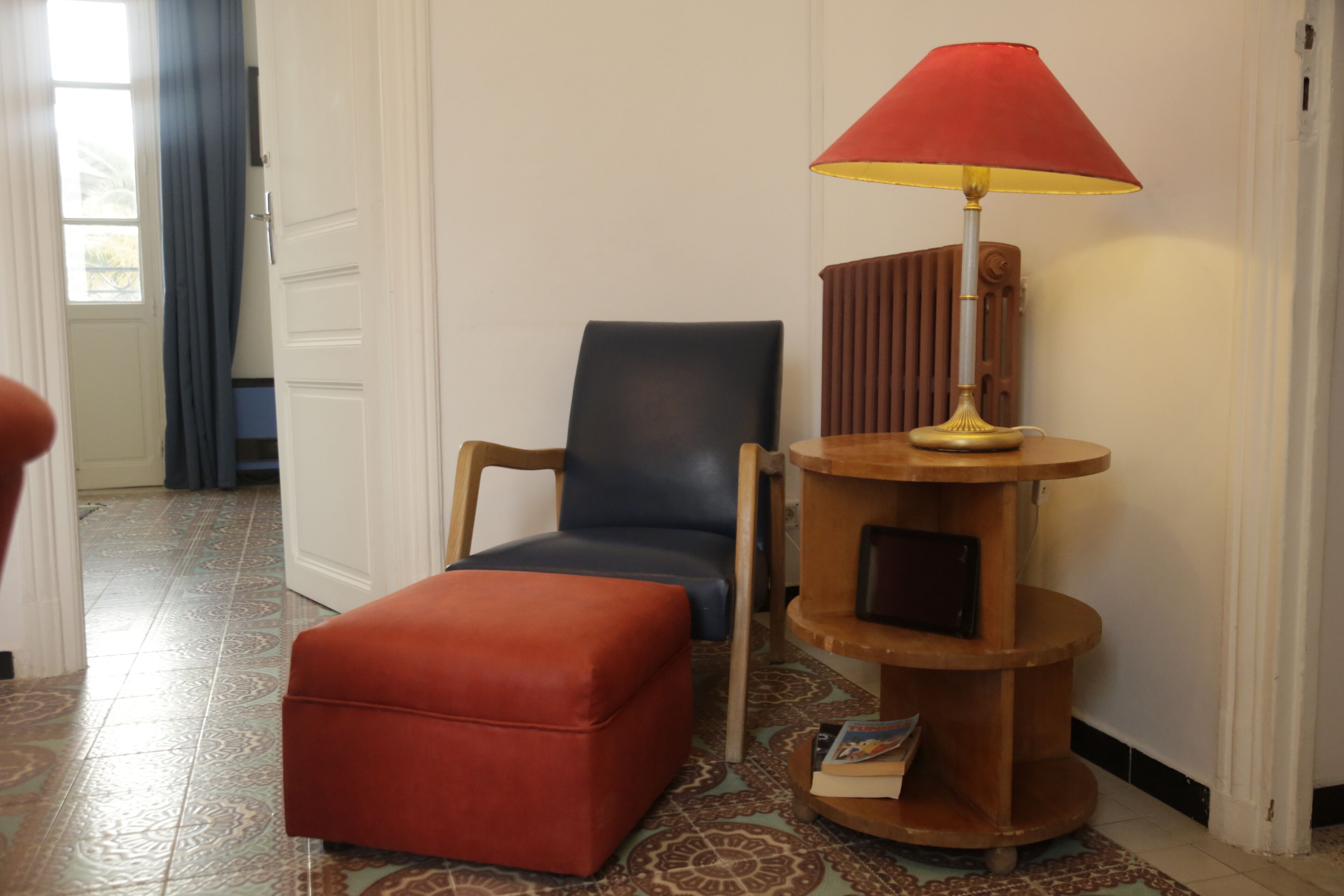 Ein Zimmer mit einem roten Hocker, einem blauen Sessel und einem Holznachttisch mit einer roten Lampe.