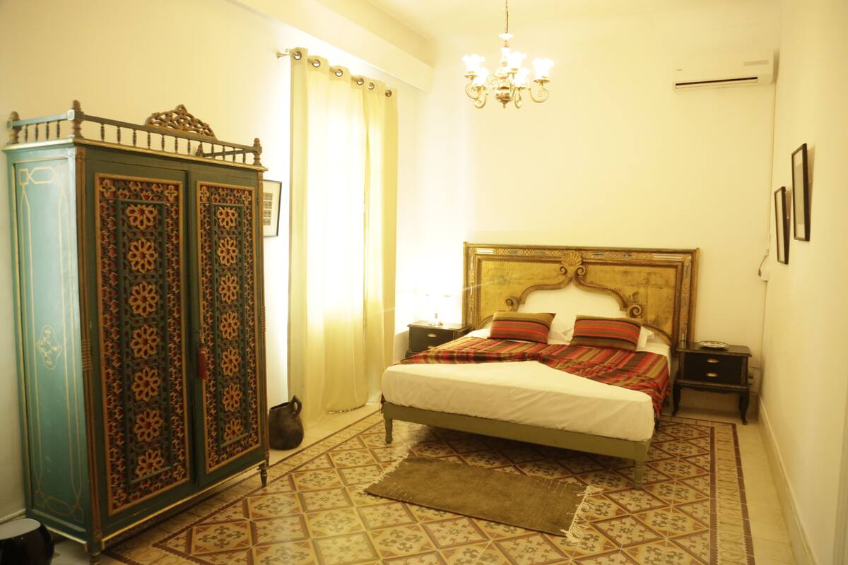 Quarto com cama, mesas de cabeceira, cômoda, lustre e ladrilhos estampados.