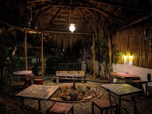 Un área de estar al aire libre con mesas y sillas, un hogar de fuego con mosaicos y una estructura cubierta de paja.