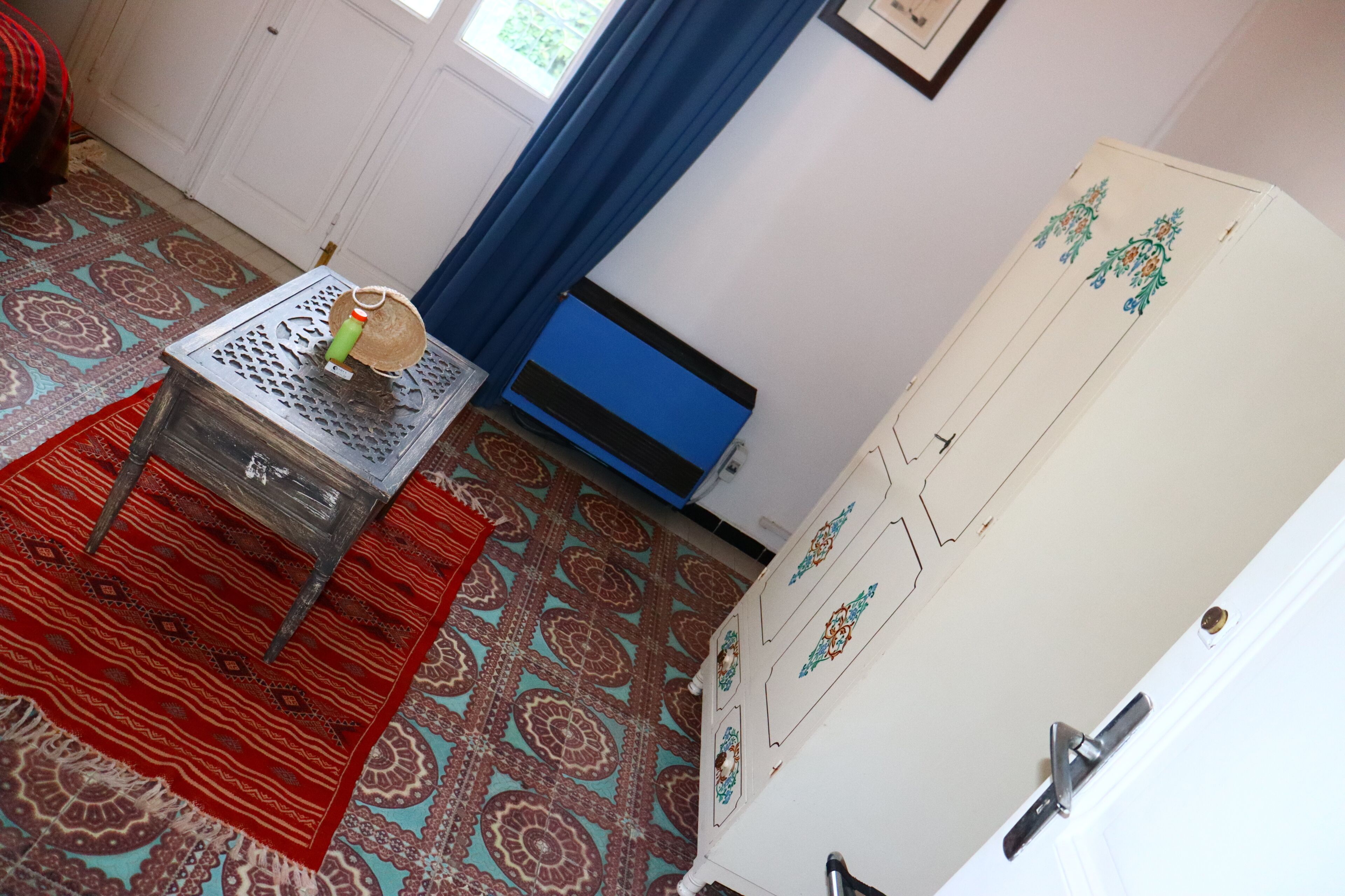 Una habitación con alfombra estampada, una mesita con una botella, una ventana con marco azul y un armario blanco con paneles decorativos.