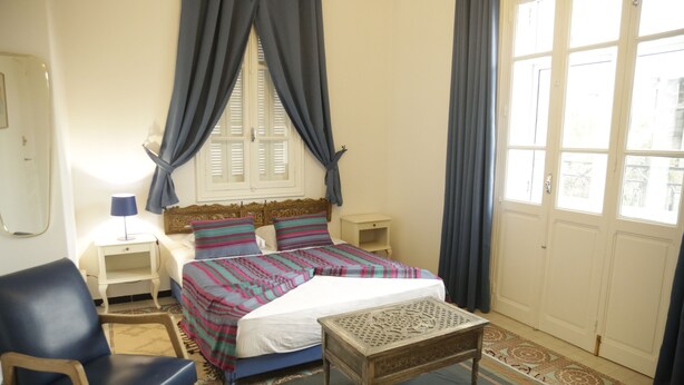 Quarto com cama, cadeira, mesa de cabeceira, luminária e janela com cortinas.