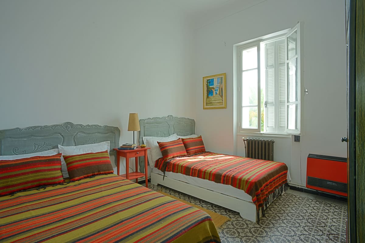 Quarto com duas camas, mesa de cabeceira, abajur, janela com persianas e radiador.
