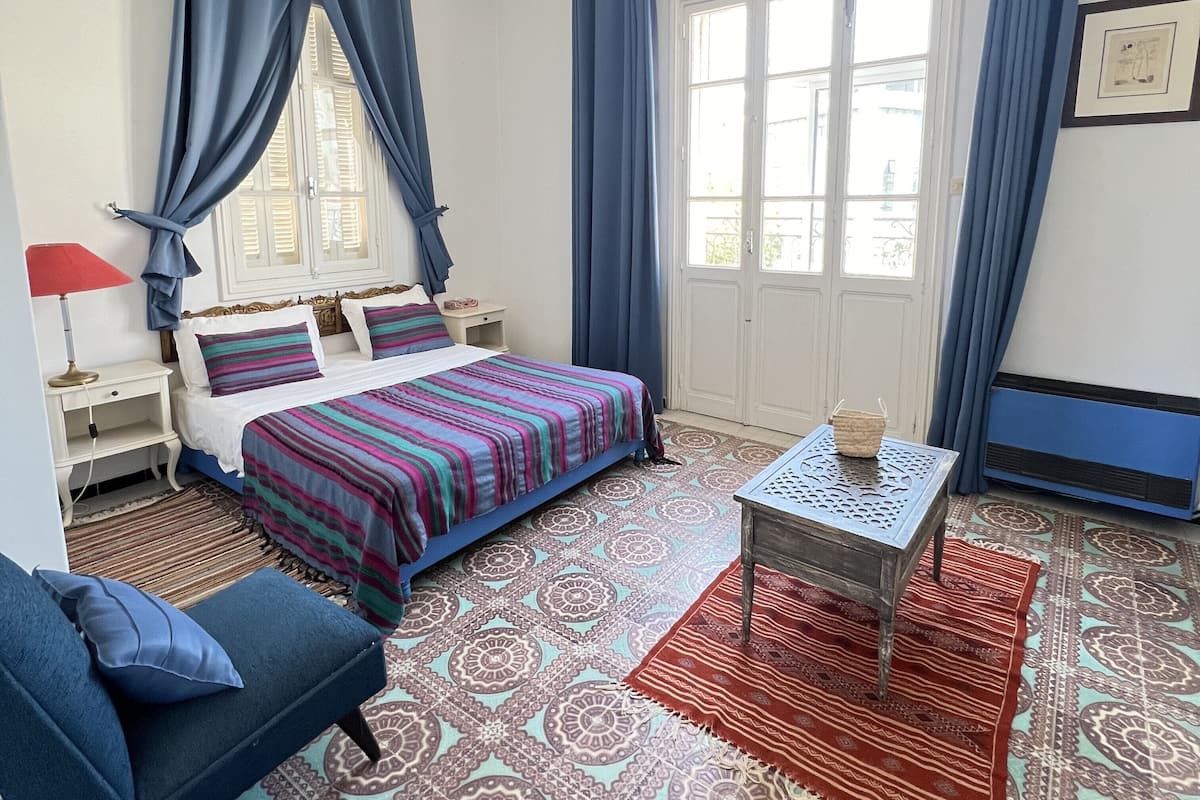 Quarto com cama, sofá azul, mesa pequena, carpete estampado e janela com cortinas.