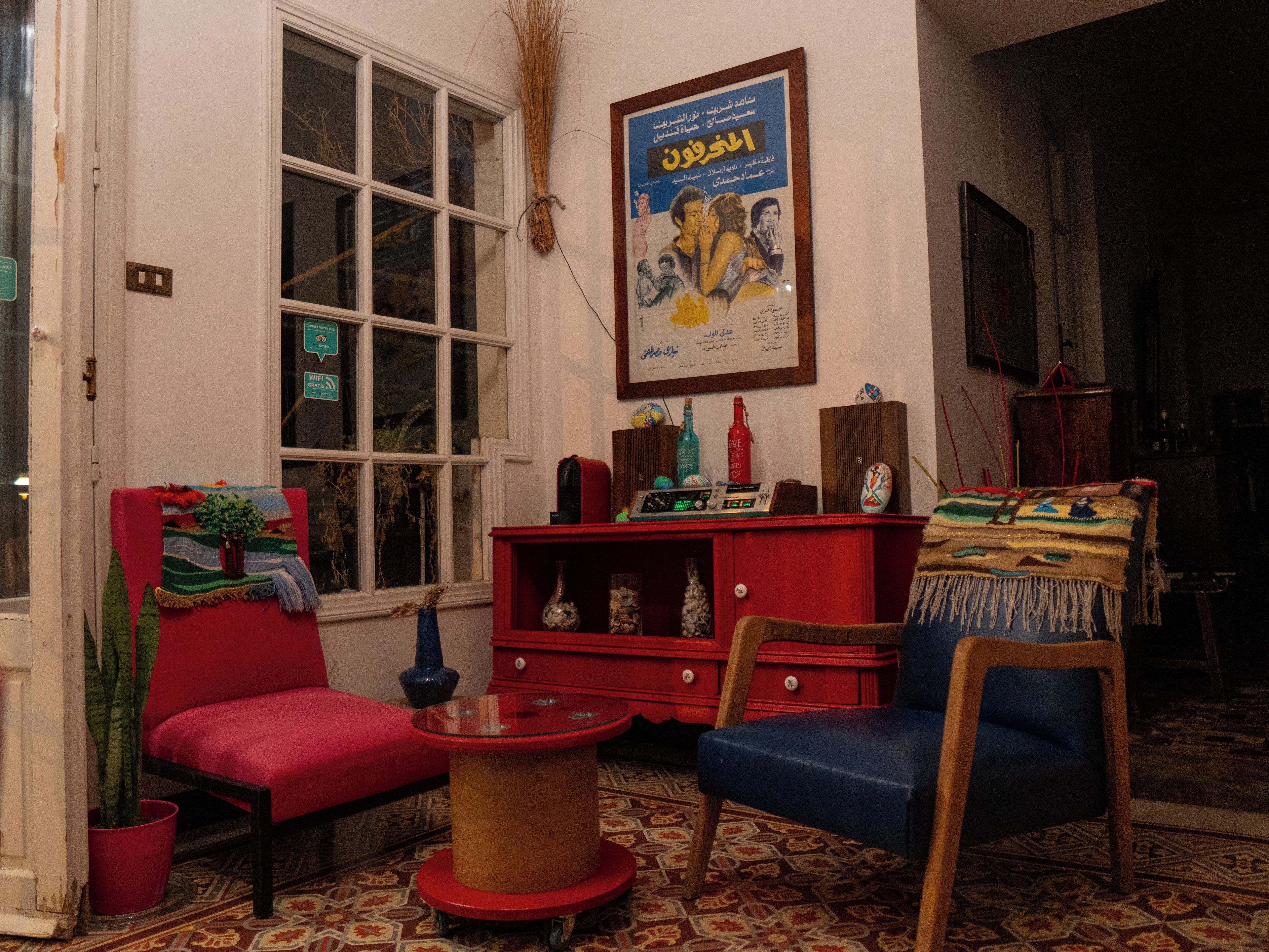 Una habitación con un sofá rojo, un sillón azul, una mesa de centro de vidrio, un armario rojo y un cartel en la pared.