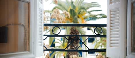 Une fenêtre ornée d’une grille en métal et de volets blancs, donnant sur un palmier et un immeuble.
