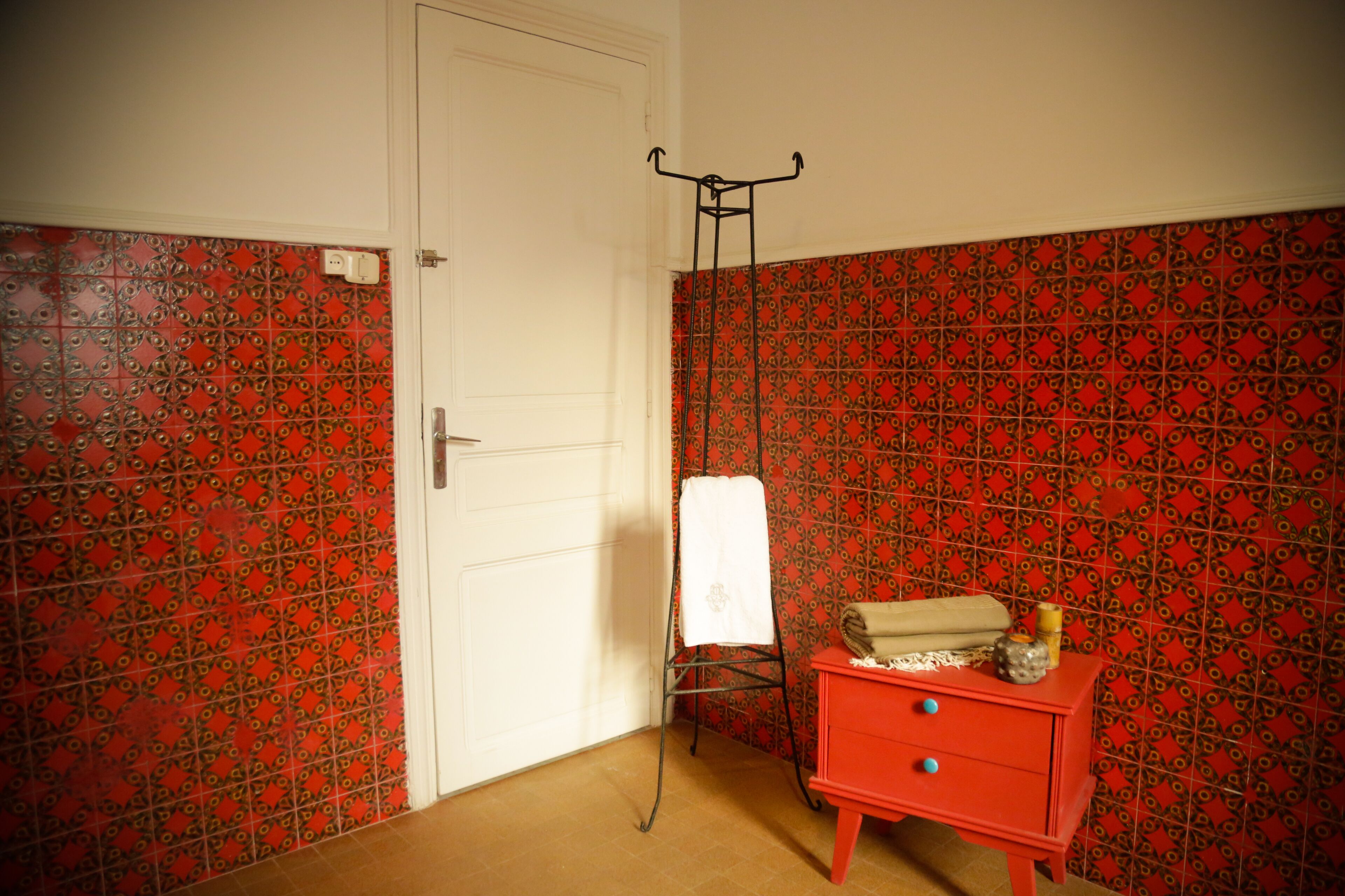 A room with patterned red and black tiles, a white door, a red dresser with drawers, and a metal drying rack with a white towel.