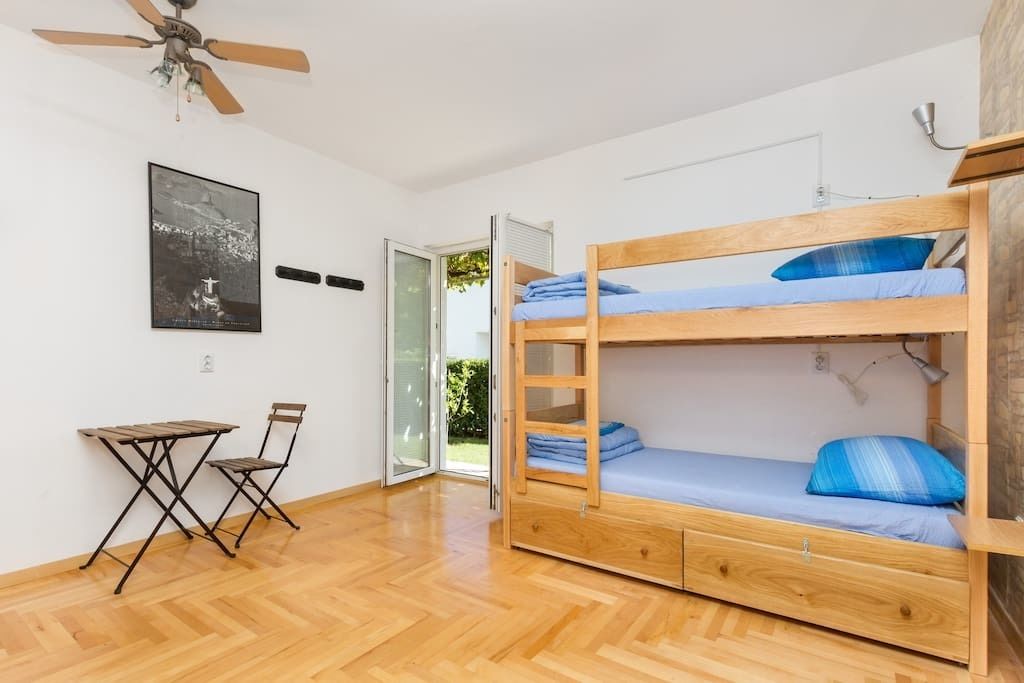 A room with a bunk bed, a small table, a chair, a ceiling fan, a door leading outside, and a framed picture on the wall.