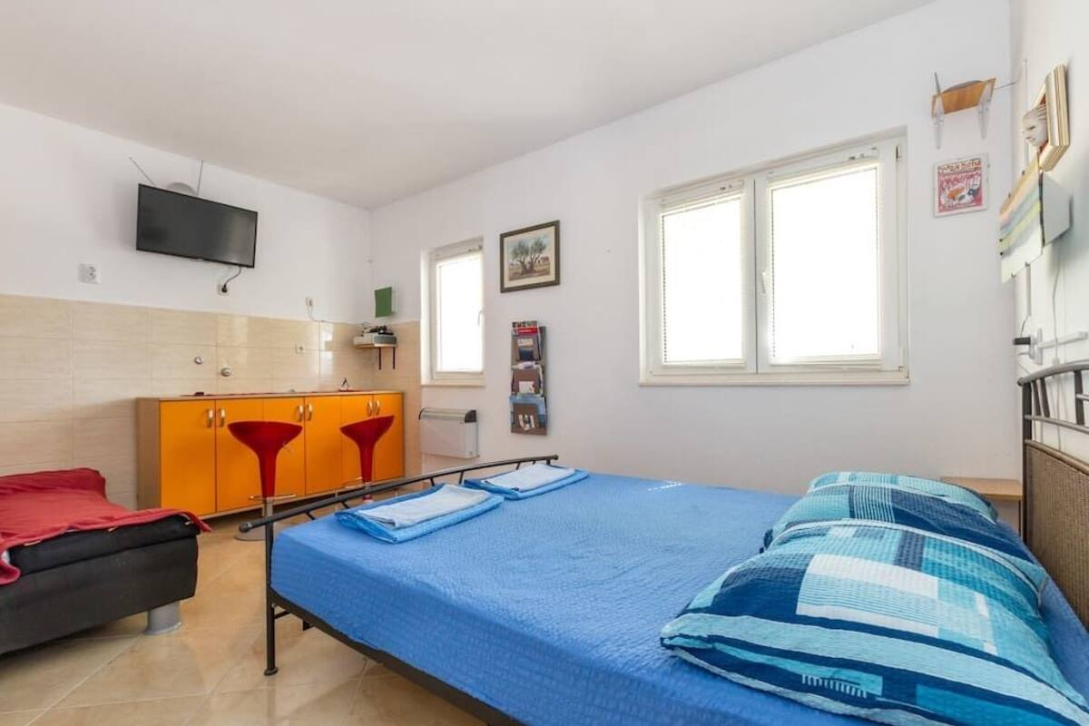 A single-bed room with a blue bedspread, a red chair, an orange cabinet, a television on the wall, and a window with blinds.