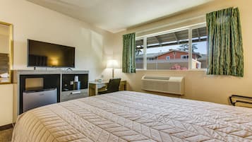 Habitación de hotel con una cama grande, un escritorio con computadora, televisión, refrigerador pequeño y una ventana con cortinas.